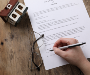 Person signing a last will and testament document beside a model house and eyeglasses, illustrating estate planning, property inheritance, and legal will preparation.
